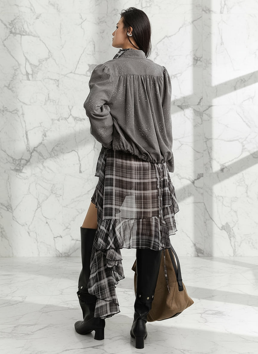 Person wearing a gray shirt and plaid skirt with black boots, standing against a white wall.